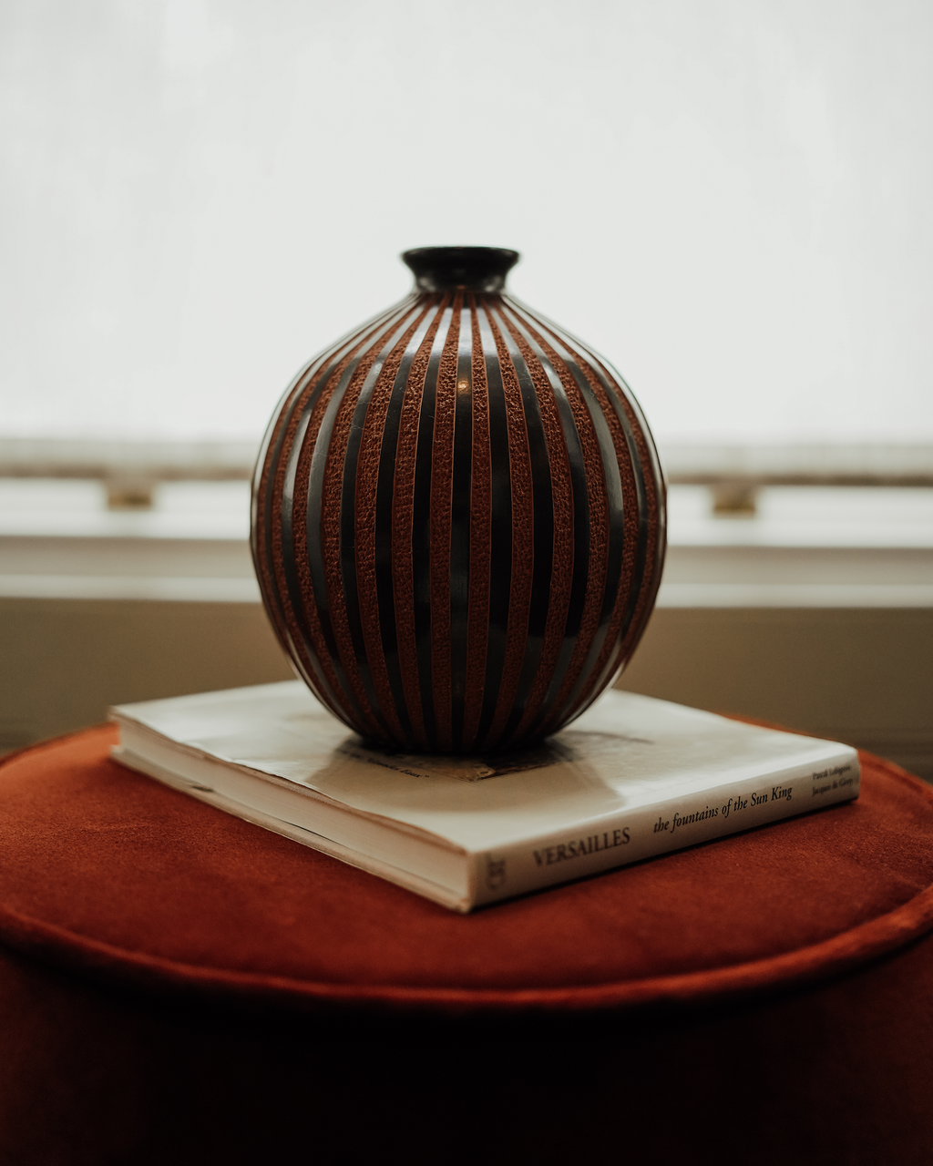 Amapola: handmade Nicaraguan ceramic round vessel with glossy black and textured terracotta vertical stripes, on a book on a red velvet stool